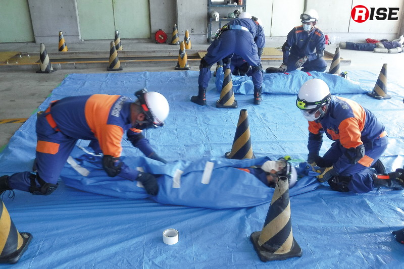 【長久手市／瓦礫救助想定】要救助者の保護と保温を目的に行うシートパッキング。三角コーンの間を活動可能範囲として訓練を行う。