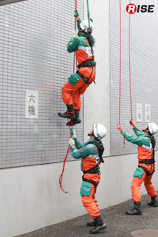 登はん員が懸垂ロープを掴むと、補助員がロープを足に巻き付ける。